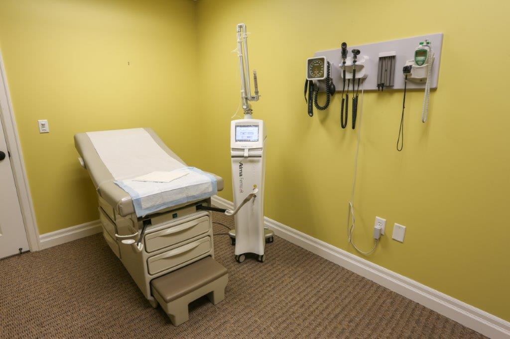 A private exam room at La Belle Vie with an exam table, wall-mounted diagnostic instruments and the Alma FemiLift laser standing beside the table