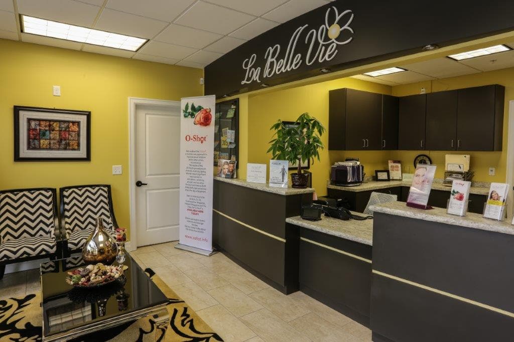 The front desk at La Belle Vie in Draper under the illuminated La Belle Vie sign, with black-and-white chevron armchairs and an O-Shot poster stand in the waiting area