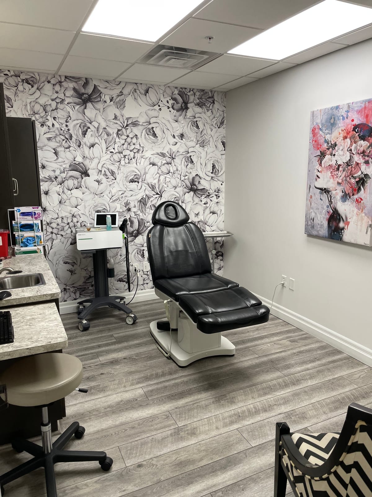 A private treatment room at La Belle Vie with a black leather treatment chair in front of a black-and-white floral feature wall, a facial device on a rolling cart, and a floral portrait painting on the grey wall