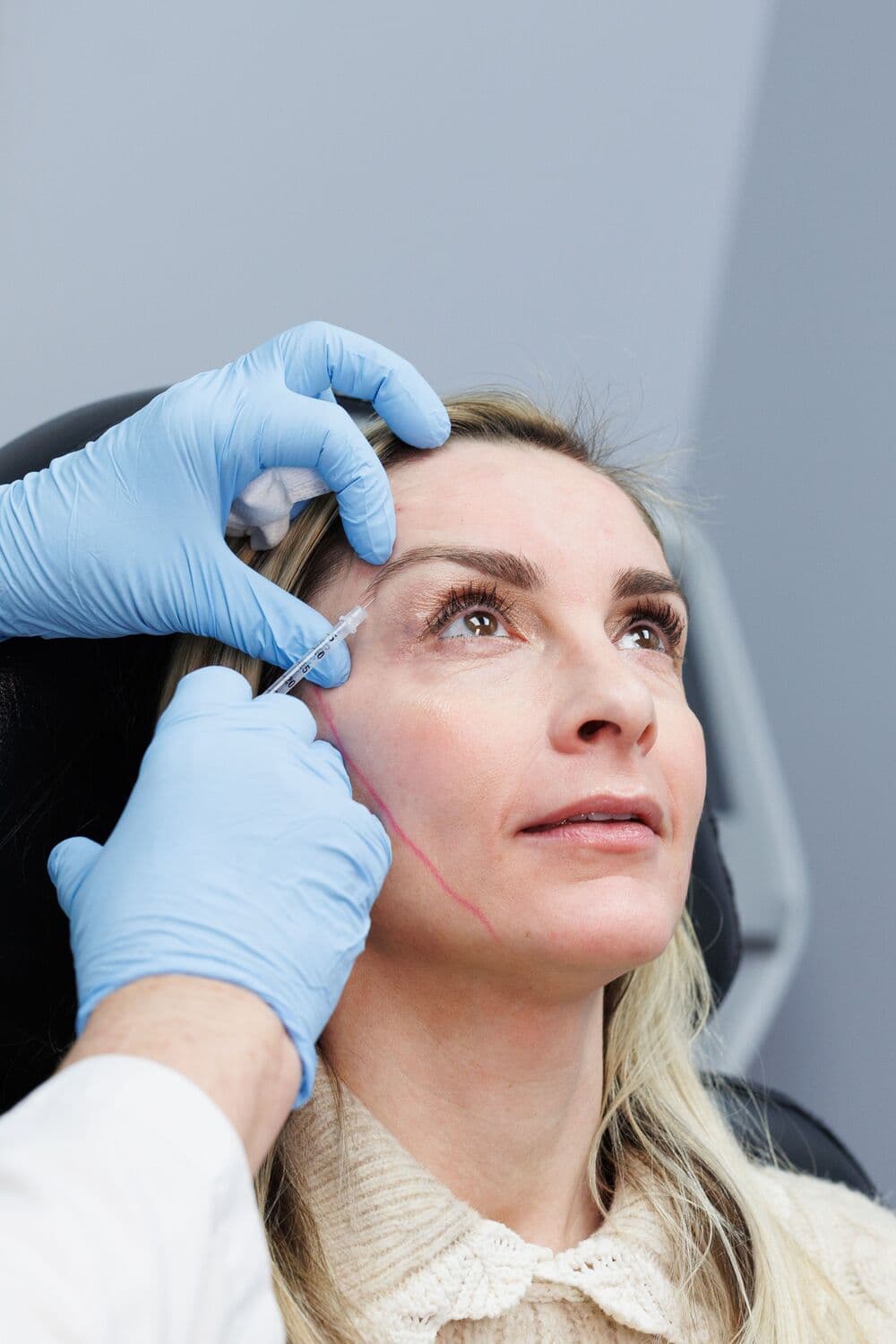 Gloved hands giving a fine-needle injection beside a blonde patient's eye at La Belle Vie, the patient looking up from the treatment chair