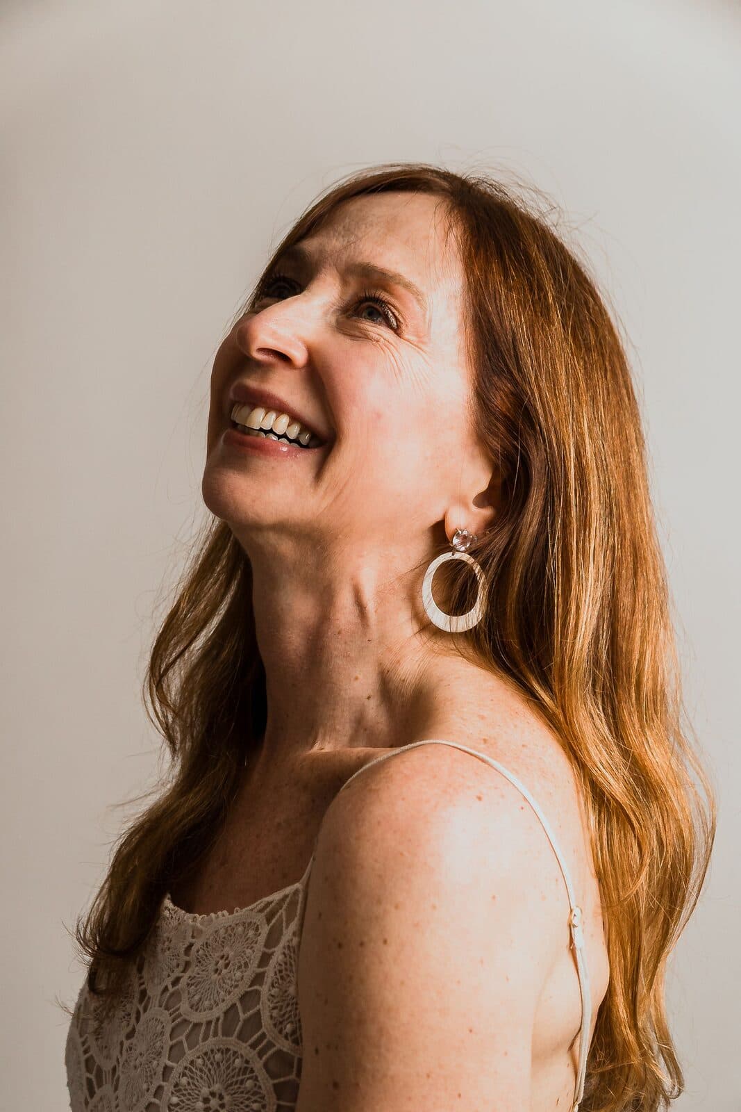 A woman in her fifties with long auburn hair laughing with her head tilted back, a hoop earring catching the light
