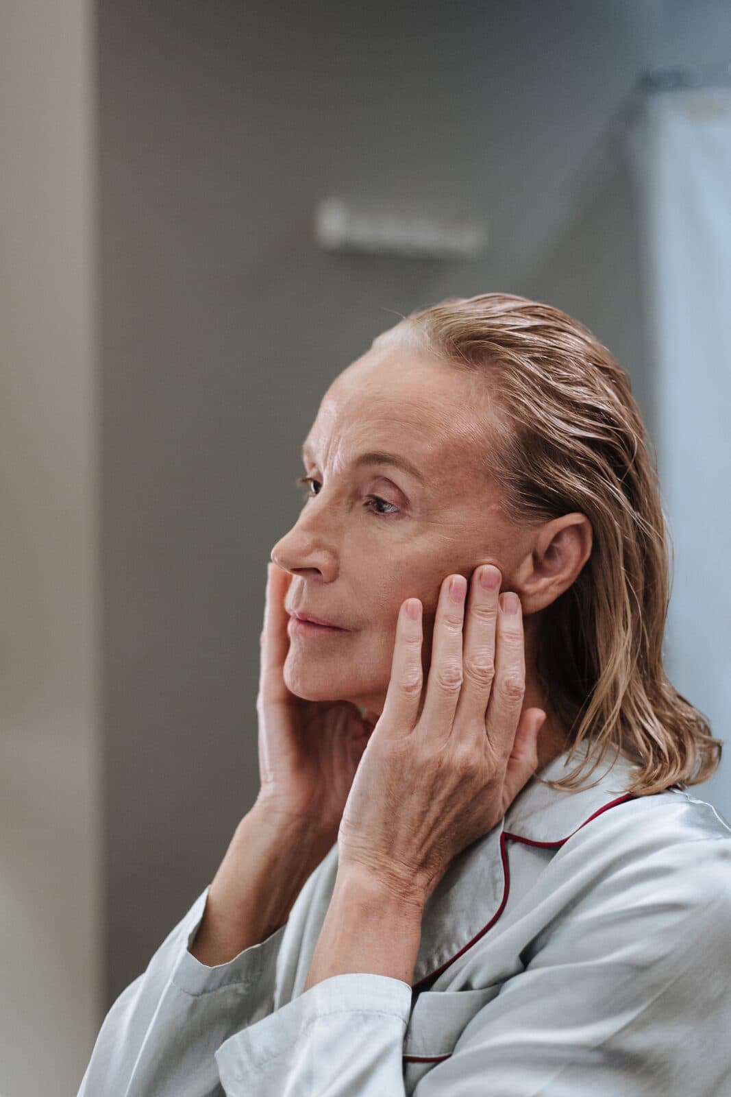 A woman in her fifties gently touching her cheeks as she checks her skin in a bathroom mirror