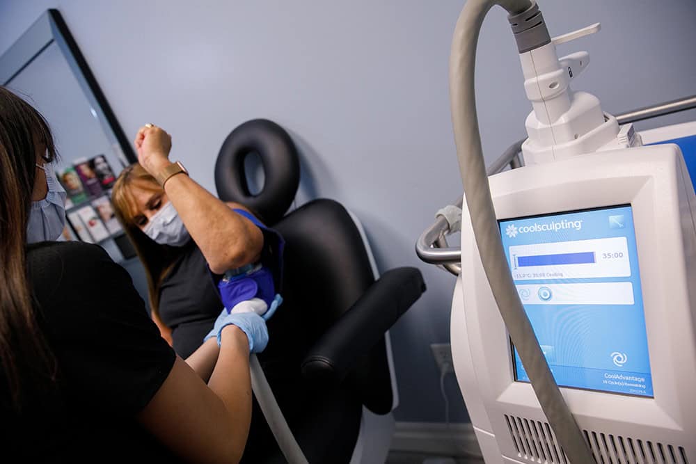 A La Belle Vie staff member in black scrubs and gloves fitting a CoolSculpting applicator to a patient's upper arm, with the CoolSculpting machine's treatment timer on screen beside the chair