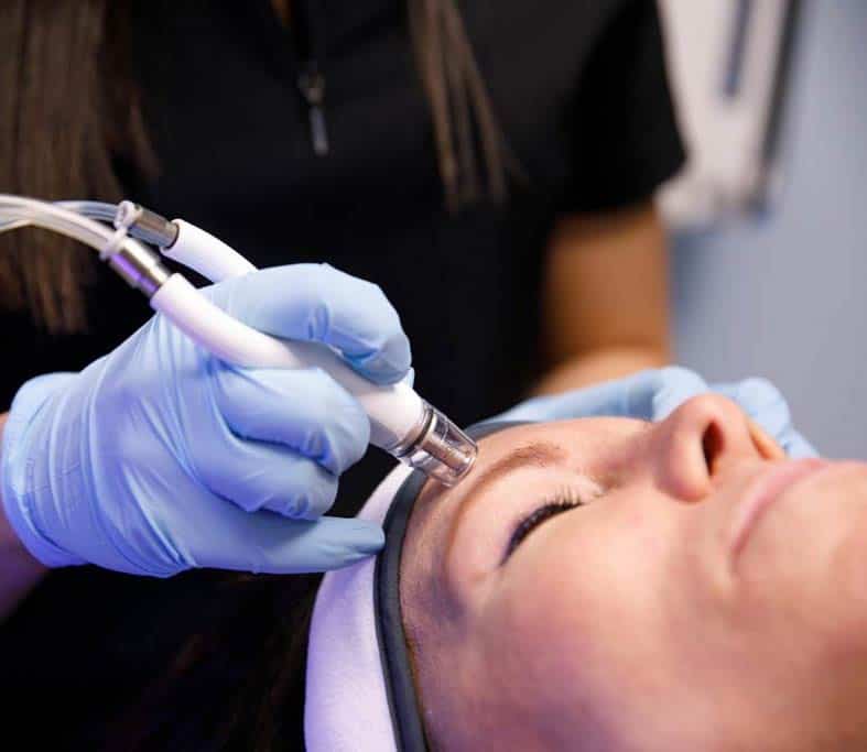 Close-up of a gloved hand gliding a DiamondGlow wand across a patient's forehead during a facial, the patient's eyes closed and hair held back by a headband