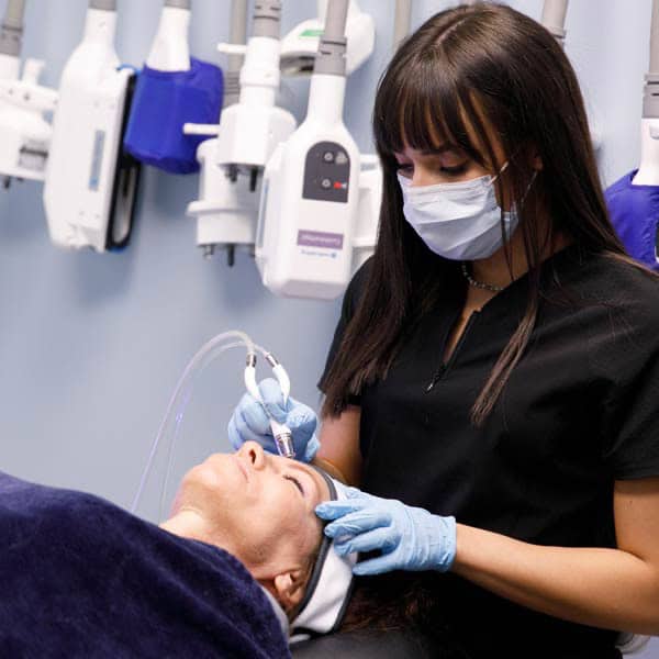 A La Belle Vie esthetician in a mask and black scrubs giving a DiamondGlow facial to a reclining patient, with CoolSculpting applicators hanging on the wall behind her