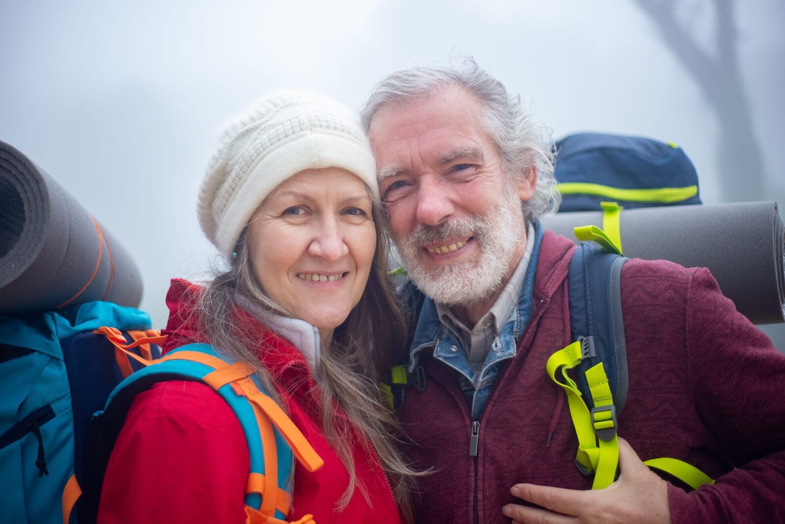 A smiling couple in their late fifties taking a selfie on a hike, backpacks on
