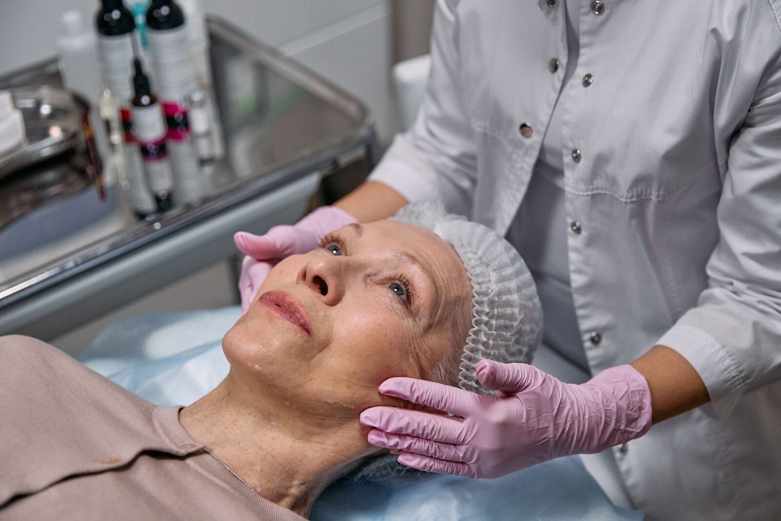 Gloved hands gently examining the facial skin of a woman in her sixties who reclines in a treatment chair