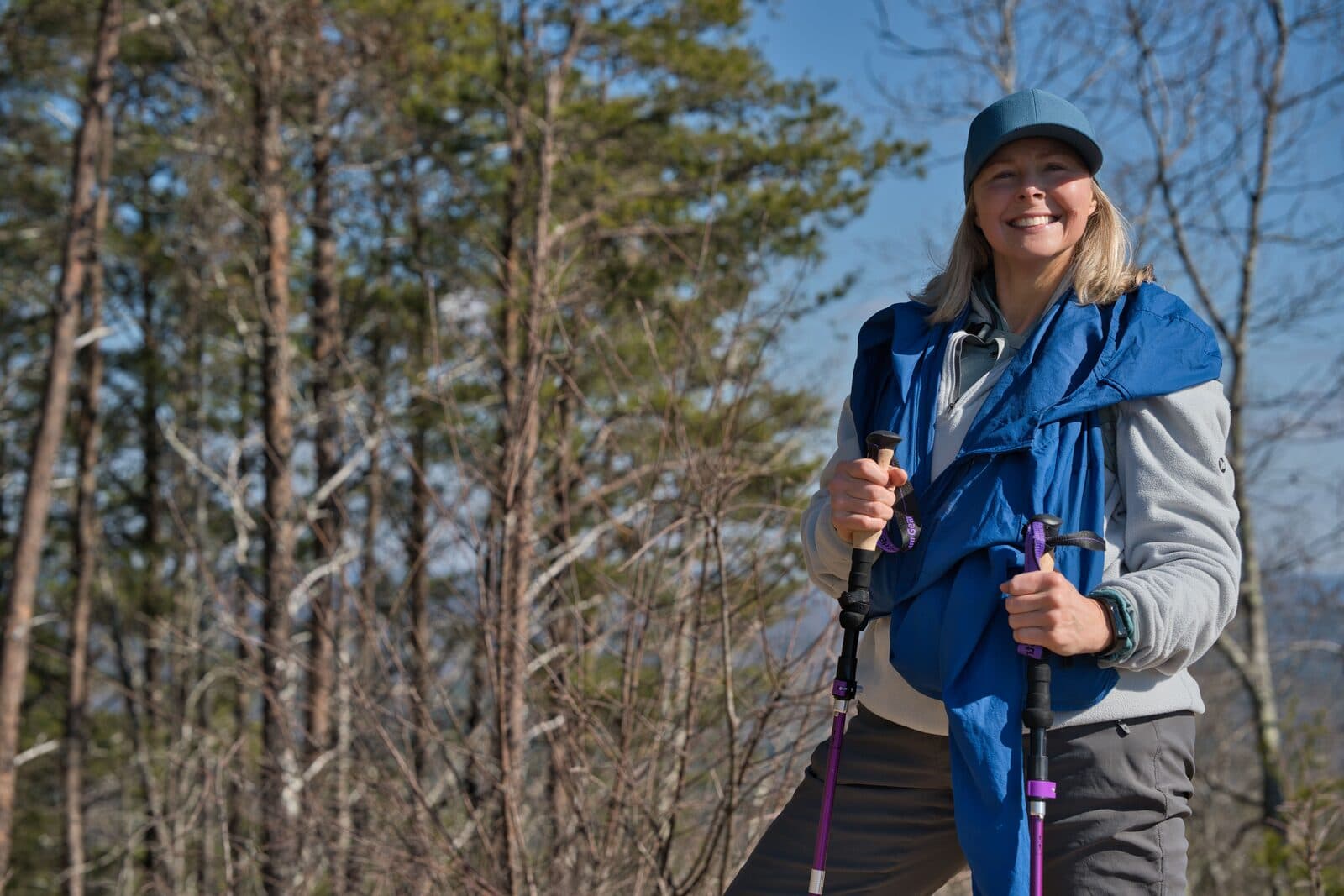 A smiling blonde woman in her fifties hiking with trekking poles among pine trees under a blue sky