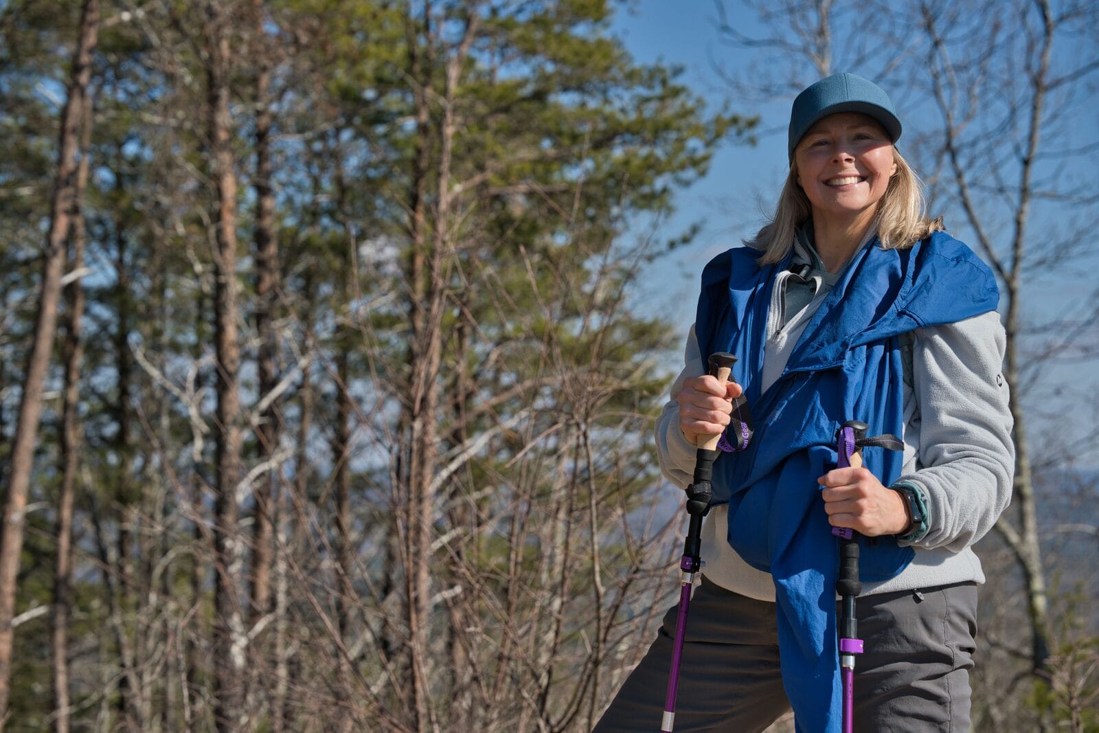 A smiling blonde woman in her fifties hiking with trekking poles among pine trees under a blue sky