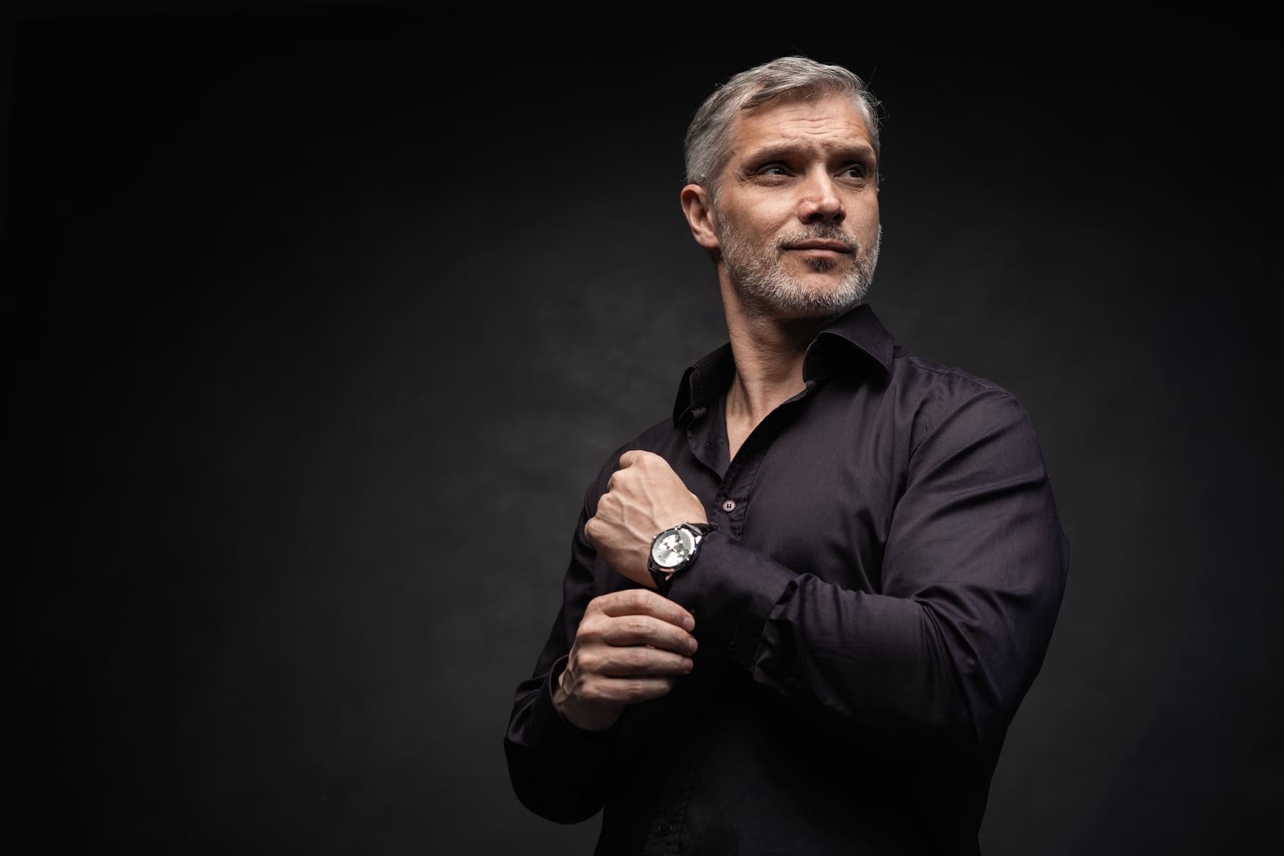 A confident gray-haired man in a dark shirt fastening his watch against a dark background