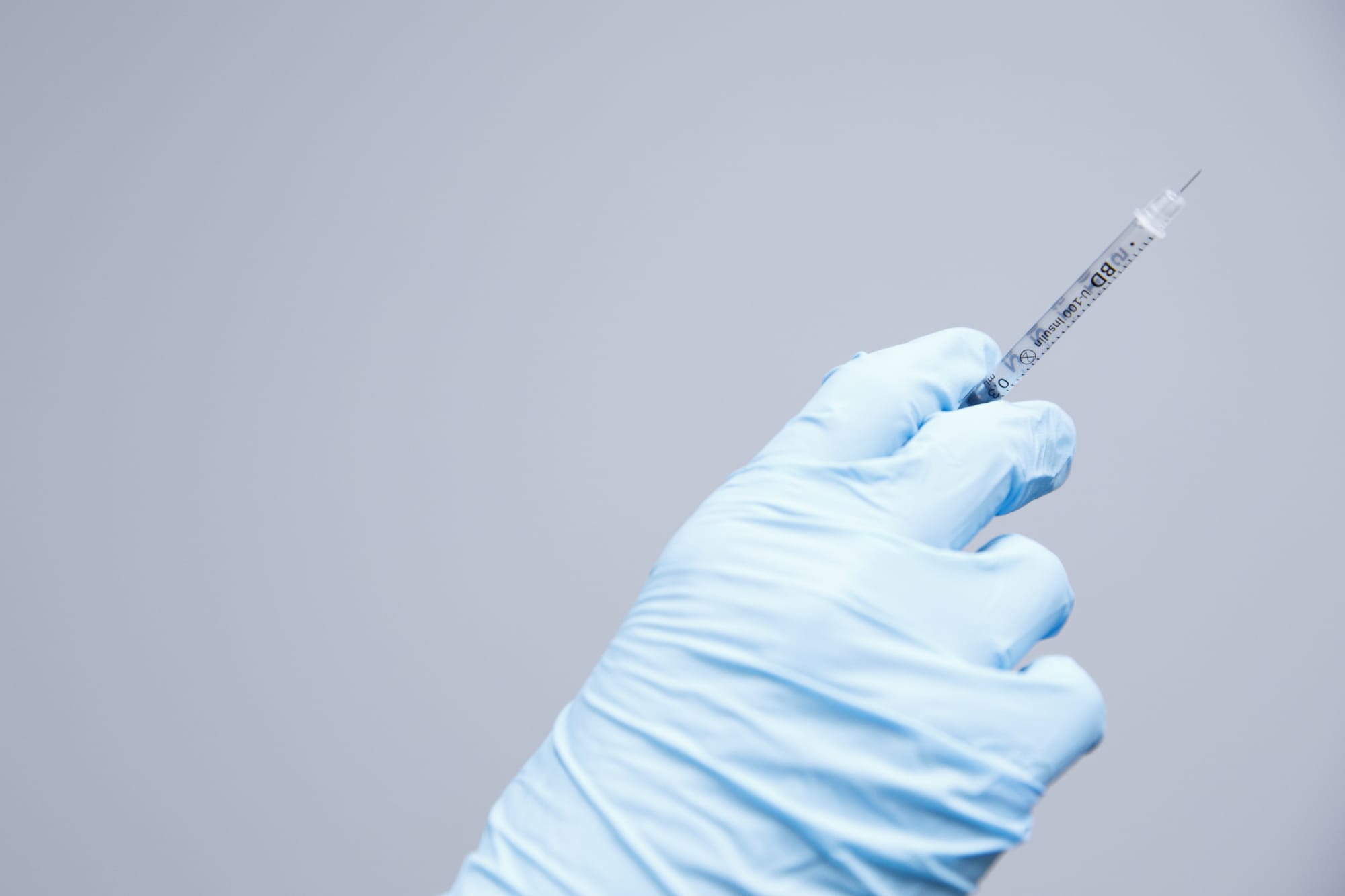 A gloved hand holding up a small syringe against a pale blue wall at La Belle Vie