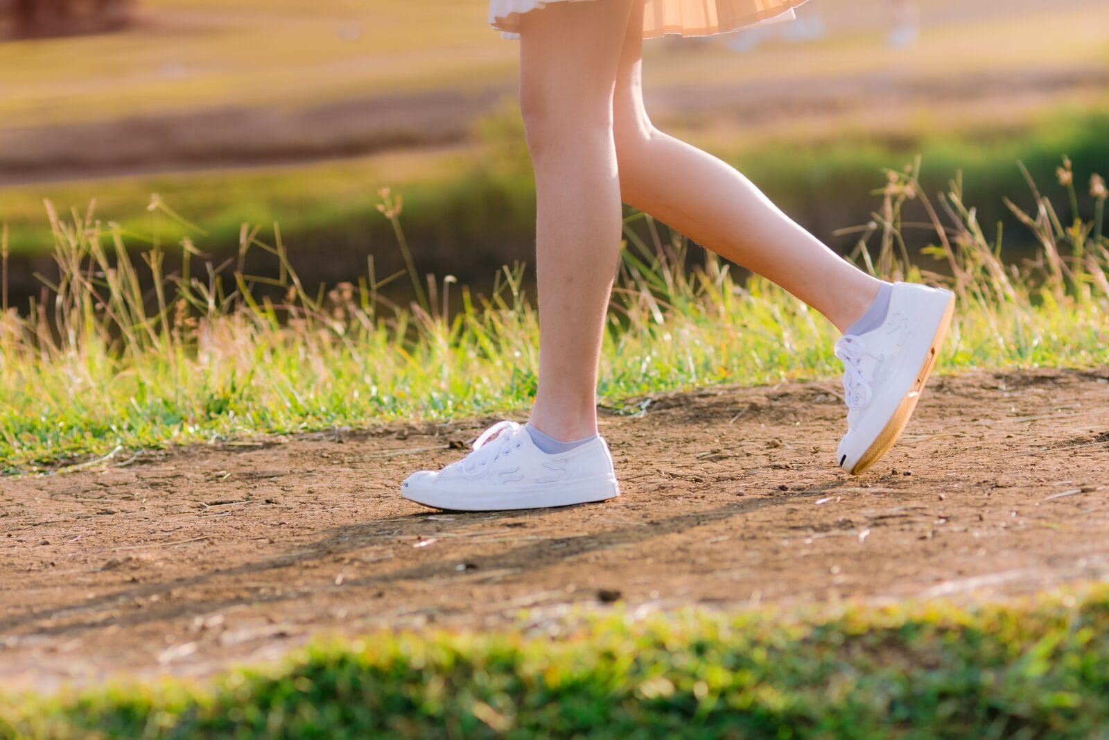 Smooth bare legs in white sneakers mid-stride on a dirt trail in summer