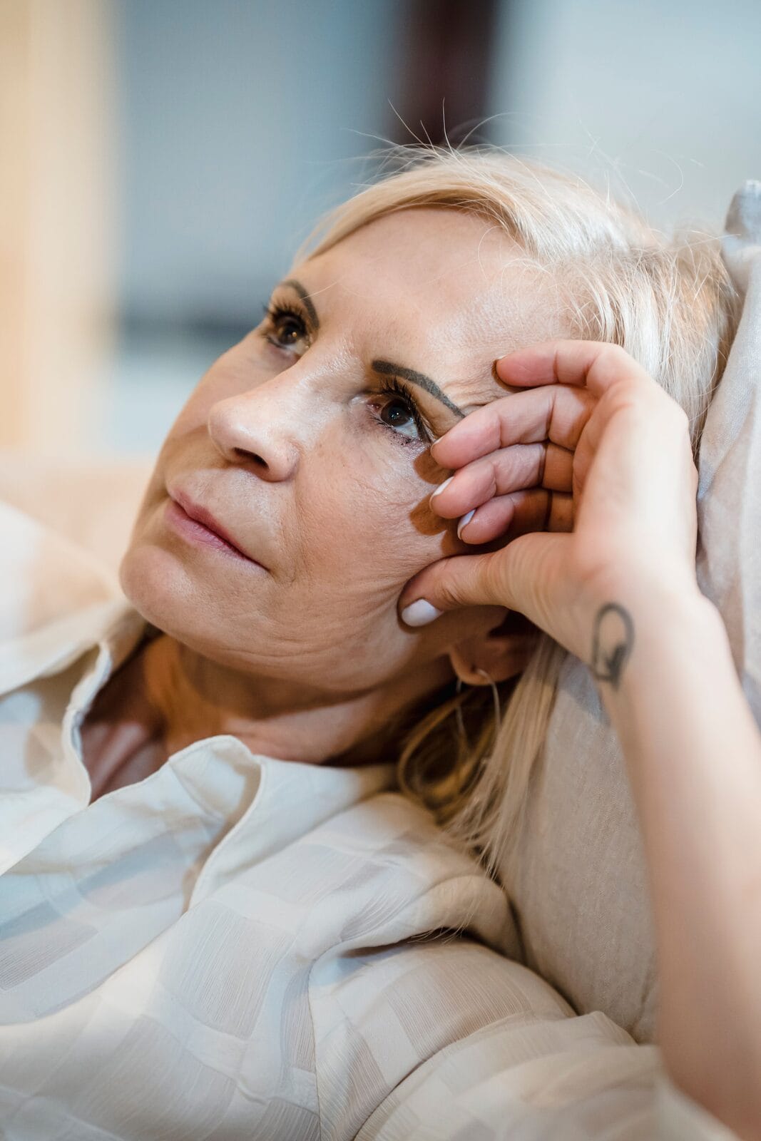A blonde woman in her fifties resting her head on a pillow, hand at her temple, looking calm and content