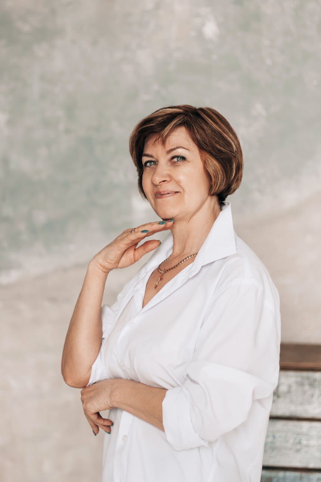 A confident woman in her fifties with short brown hair in a white shirt, one hand resting at her collarbone