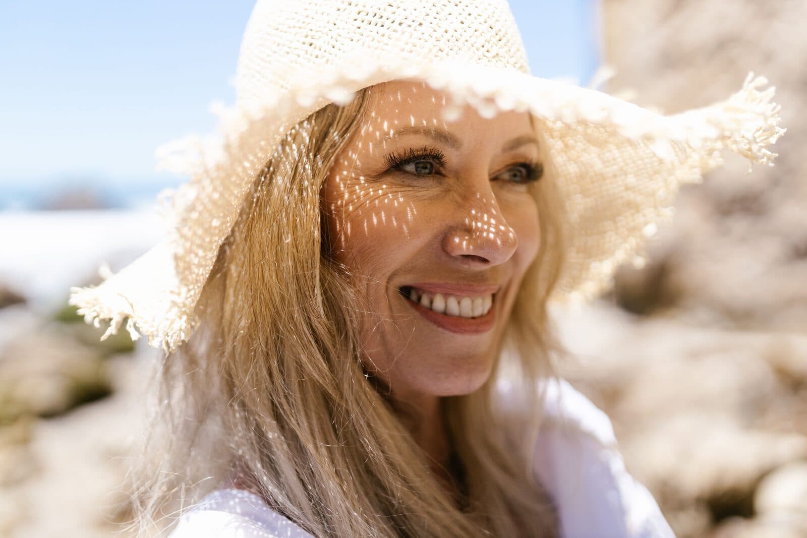 A blonde woman in her fifties laughing in a straw sun hat, sunlight filtering across her face